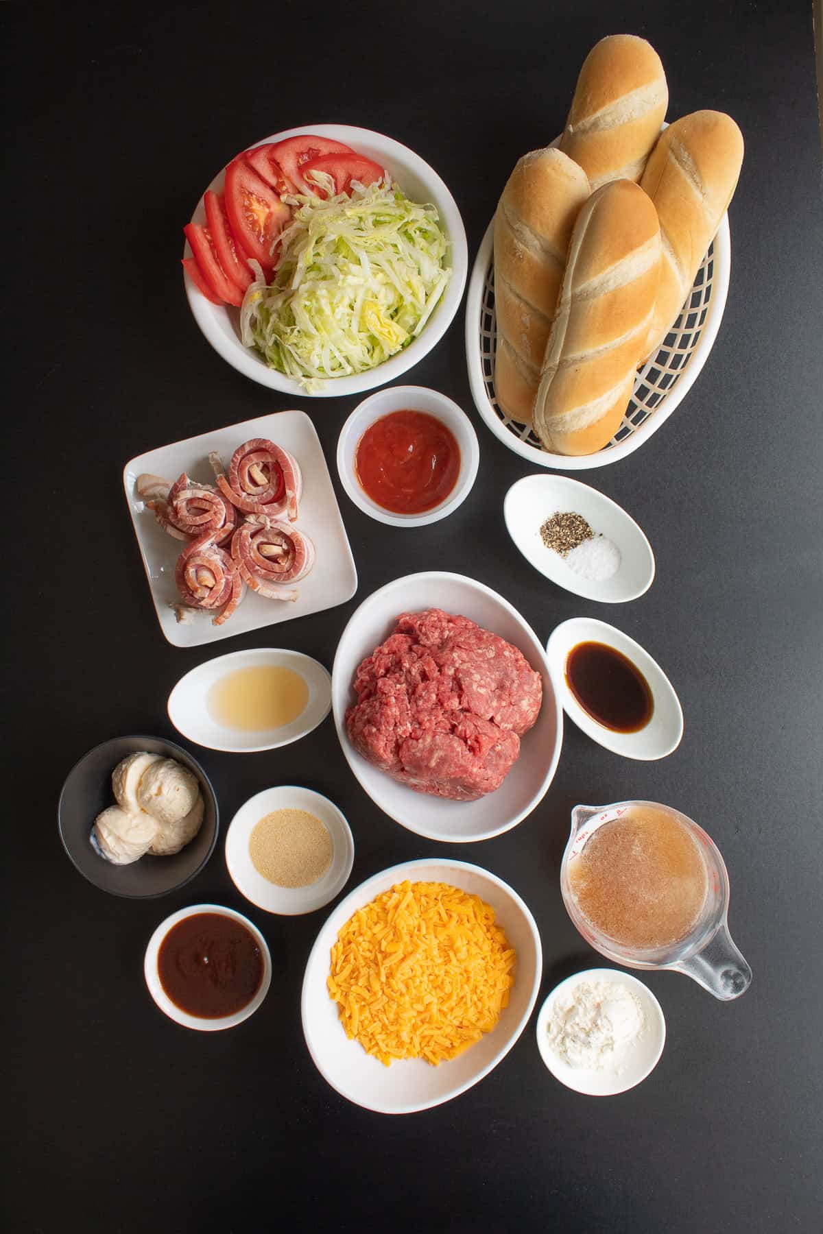 The ingredients for the cheeseburger subs are arranged in small bowls and plates on a black surface.