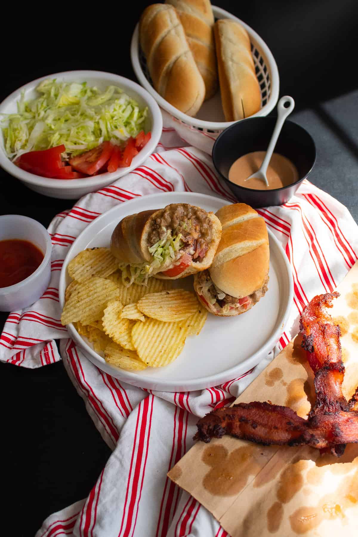 A bacon cheeseburger sub topped with lettuce and special sauce on a plate alongside potato chips and extra toppings.