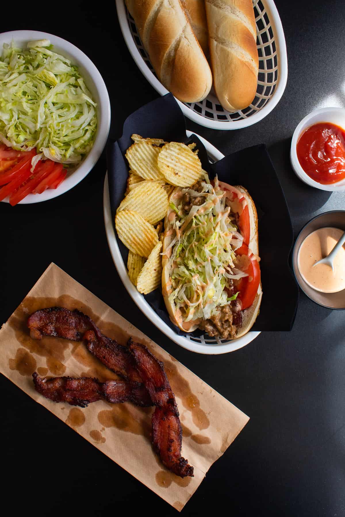 A bacon cheeseburger sub topped with lettuce and special sauce in a basket alongside potato chips and extra toppings.