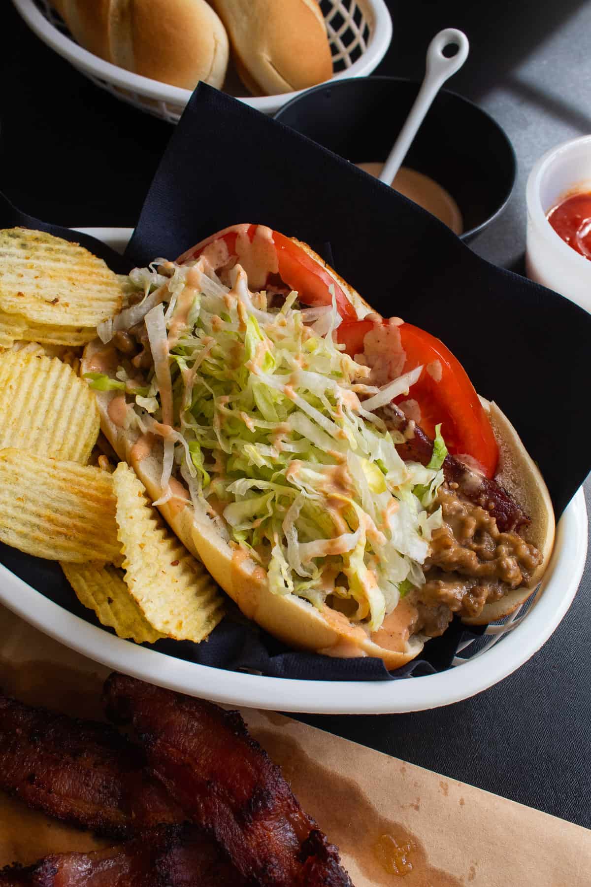 A bacon cheeseburger sub topped with lettuce and special sauce in a basket alongside potato chips.