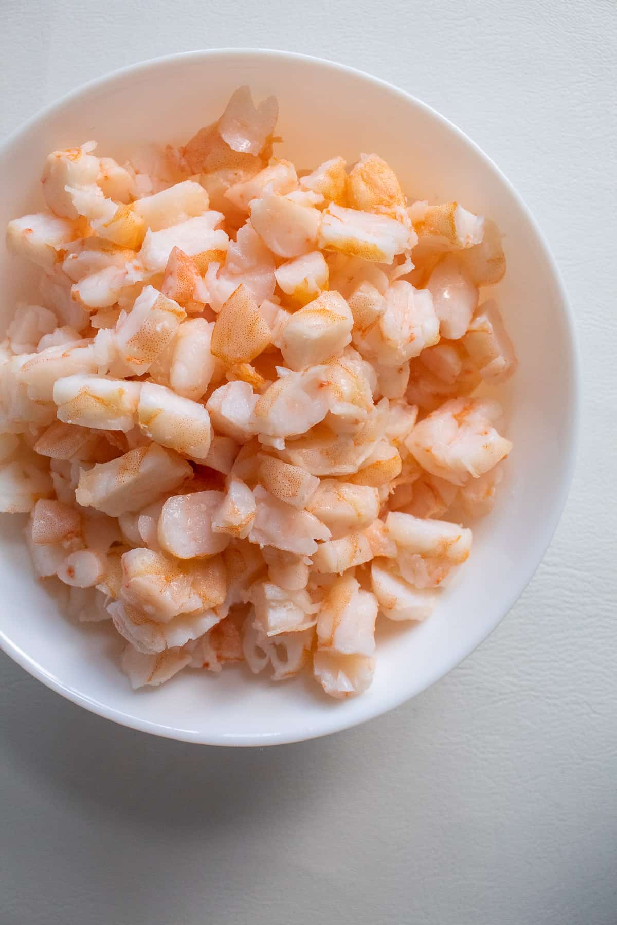 A white bowl containing chopped cooked shrimp sits on a white surface.