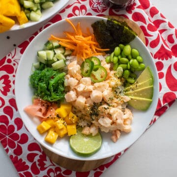 A white bowl containing shrimp poke and a variety of toppings sits on top of a red and white patterned cloth.