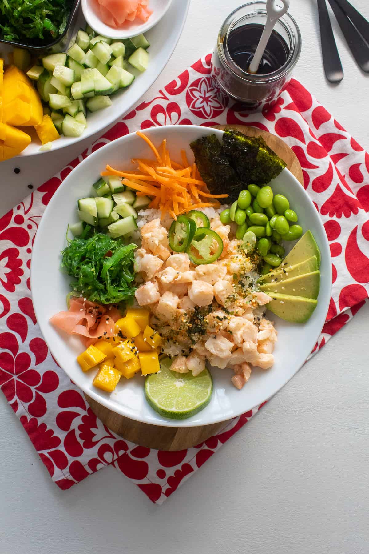 A white bowl containing shrimp poke and a variety of toppings sits on top of a red and white patterned cloth alongside a jar of dark sauce and a platter of additional toppings.