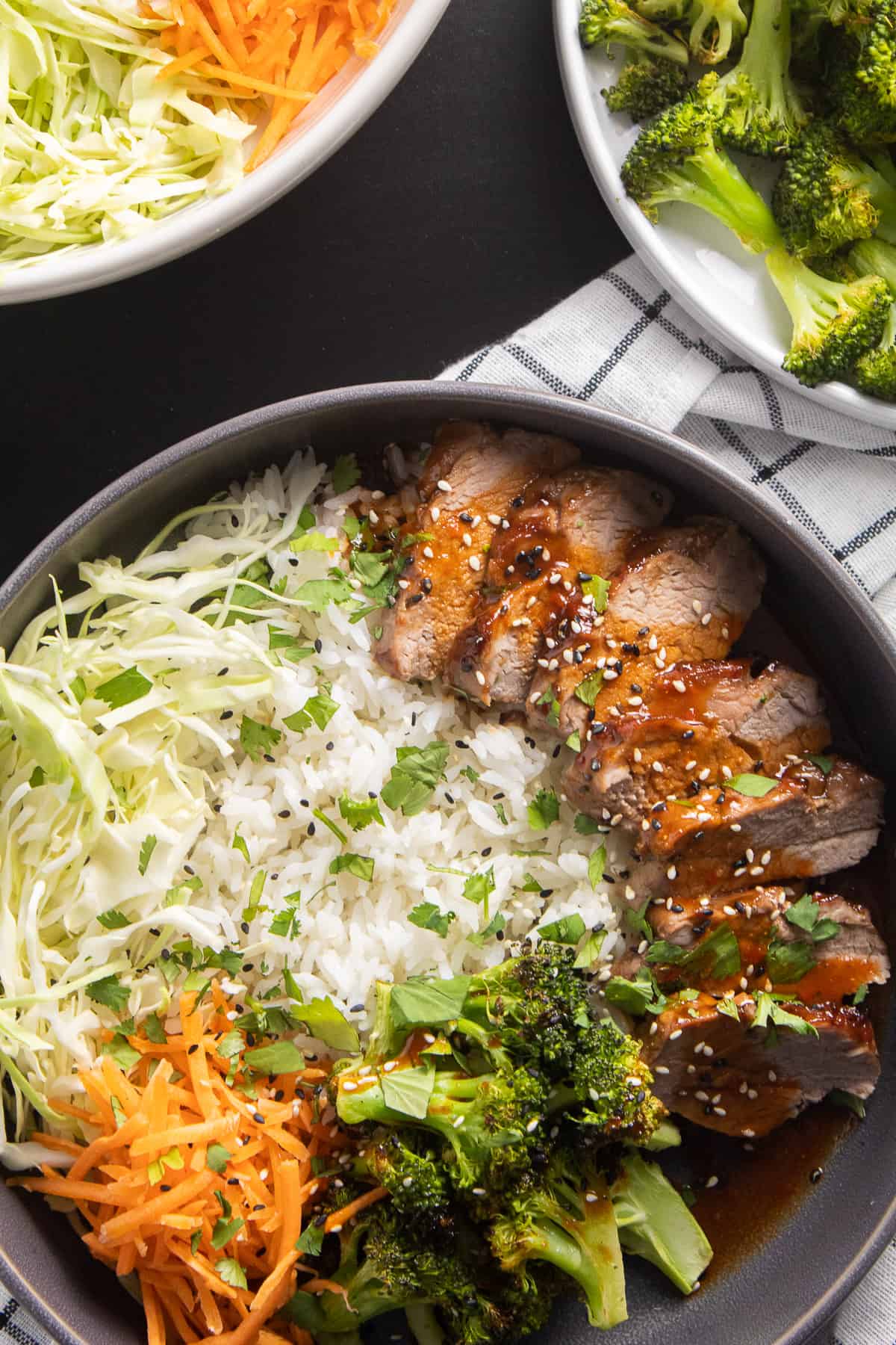 Pork tenderloin slices sit on a bed of rice alongside roasted broccoli, raw carrots, and cabbage and are drizzled with a orange-red sauce and sprinkled with sesame seeds and cilantro.