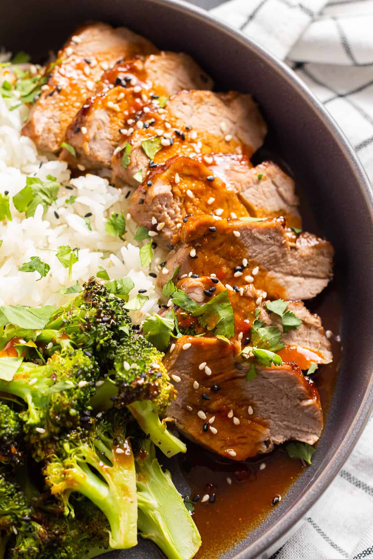 Pork tenderloin slices sit on a bed of rice alongside roasted broccoli and are drizzled with a orange-red sauce and sprinkled with sesame seeds and cilantro.
