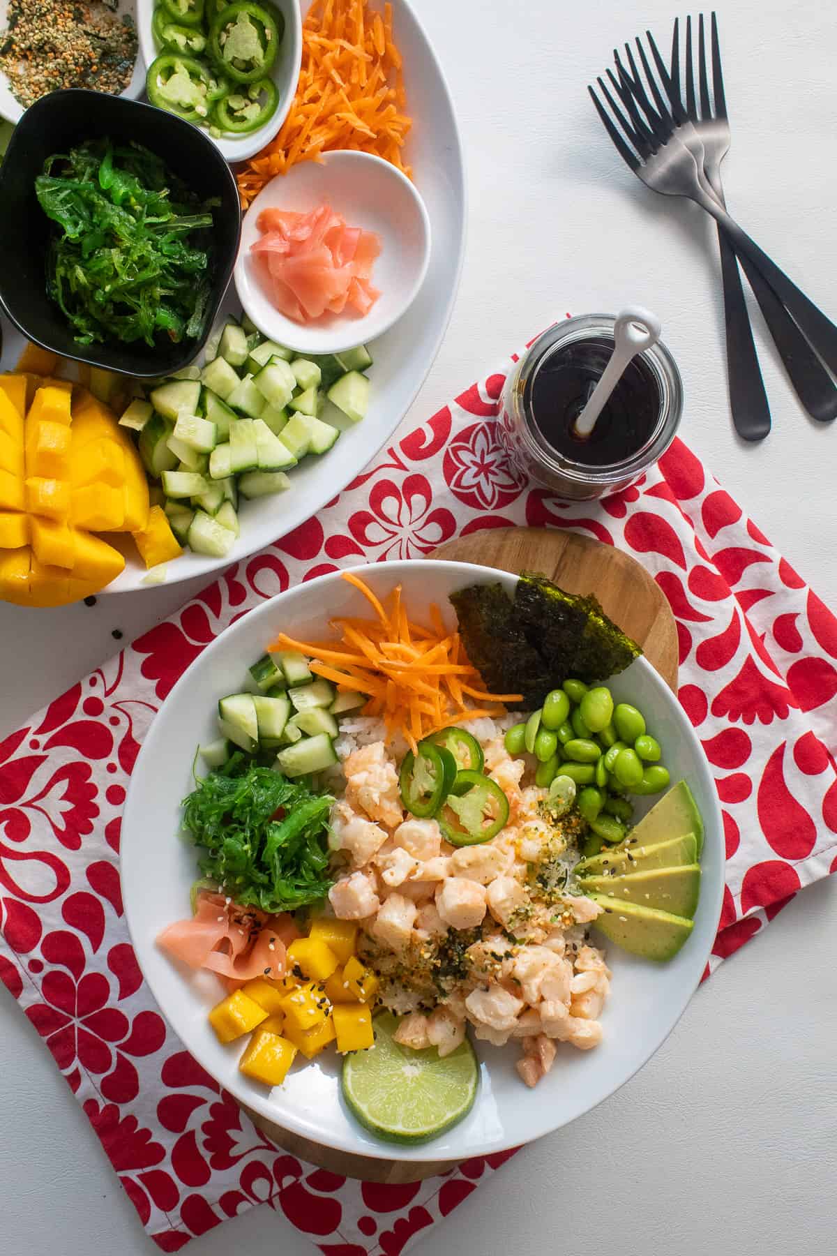 A white bowl containing shrimp poke and a variety of toppings sits on top of a red and white patterned cloth alongside a jar of dark sauce and a platter of additional toppings.