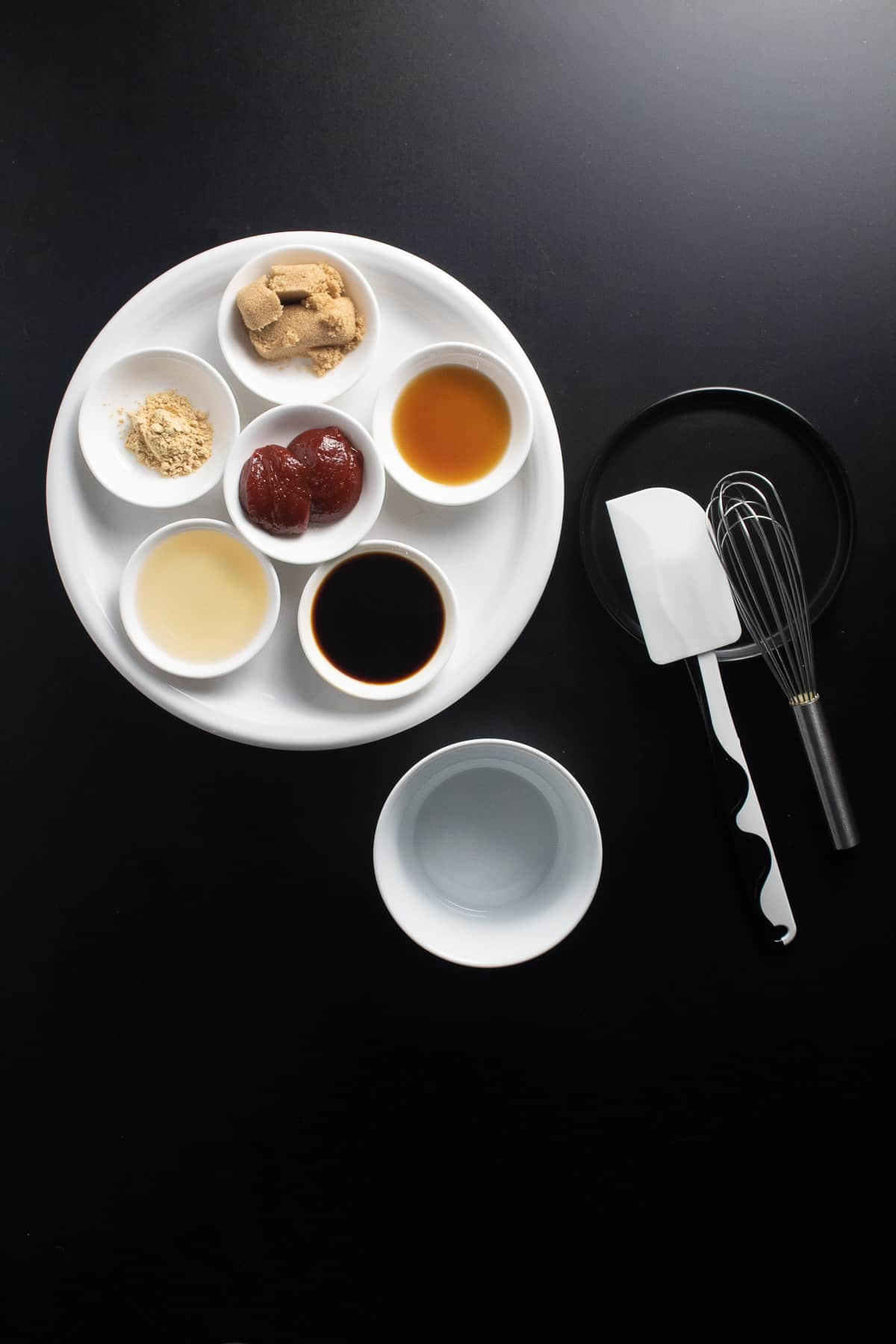 Ingredients for the gochujang sauce are arranged on a white plate on a black surface and next to a spatula, whisk, and small bowl.