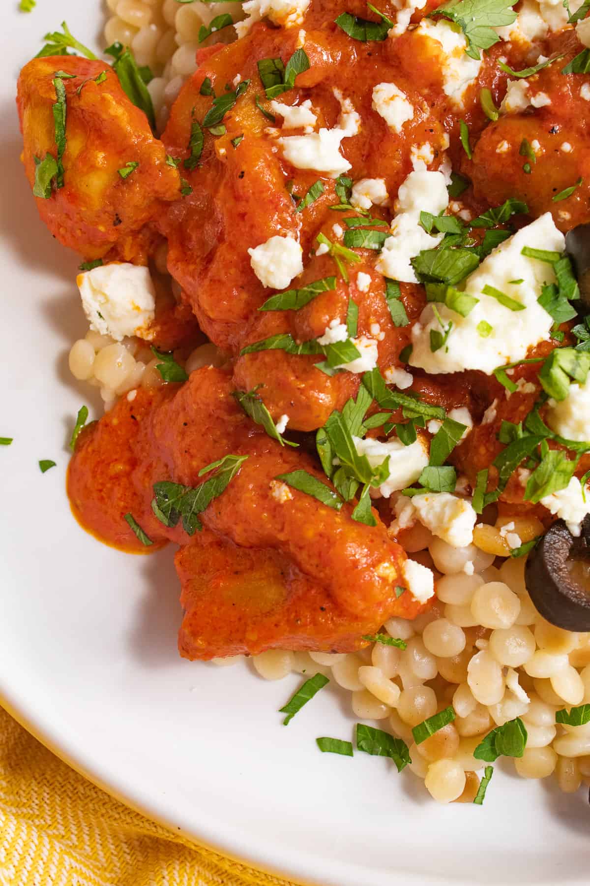 A detailed close-up of the orange sauce, chicken, parsley, feta, and olives piled on a white plate.