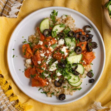 A white plate is piled with pearl couscous, a vibrant orange sauce, and cucumber, feta cheese, parsley, and olive garnishes.