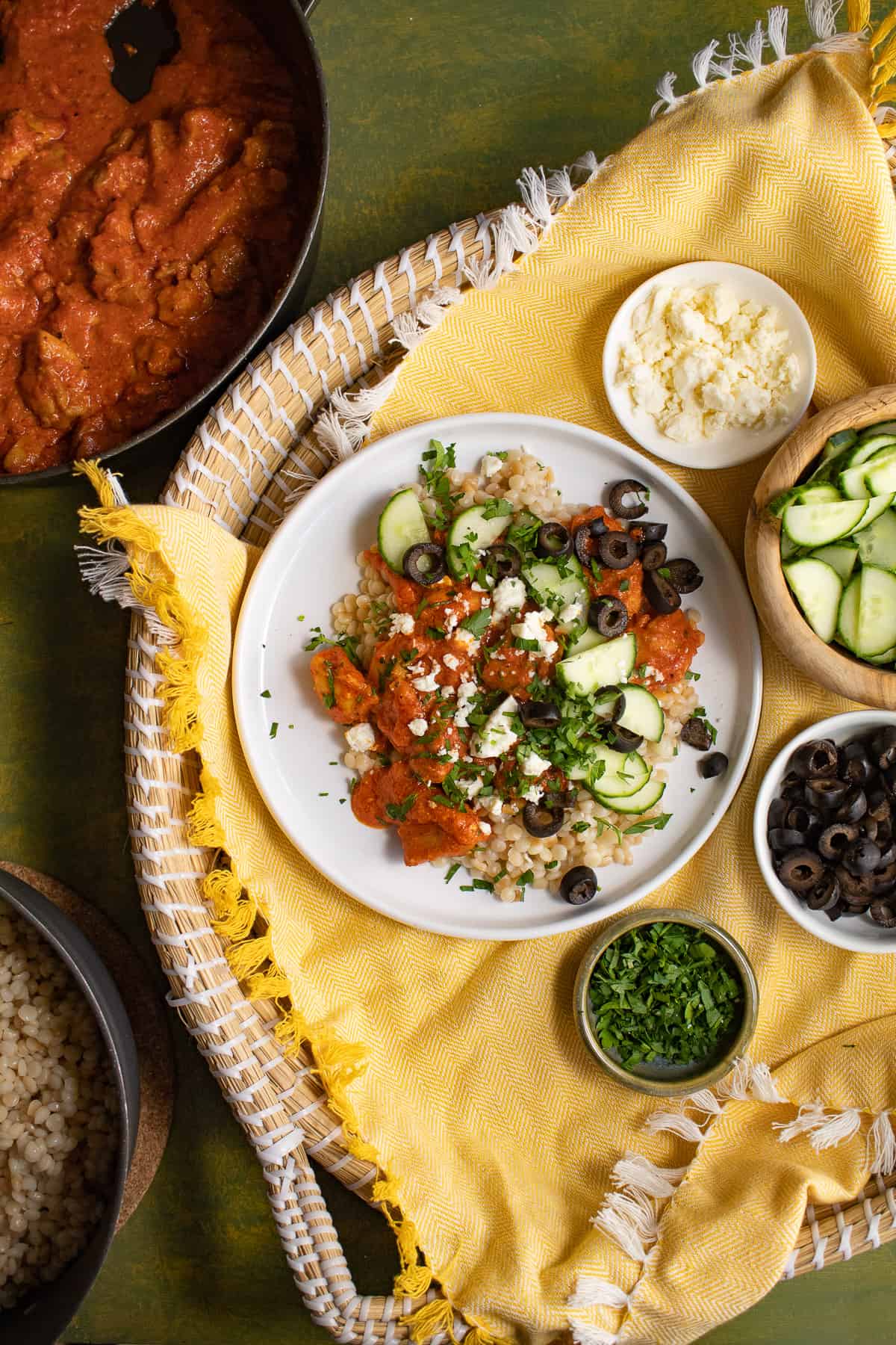 A white plate is piled with pearl cous cous, a vibrant orange sauce, and cucumber, feta cheese, parsley, and olive garnishes.