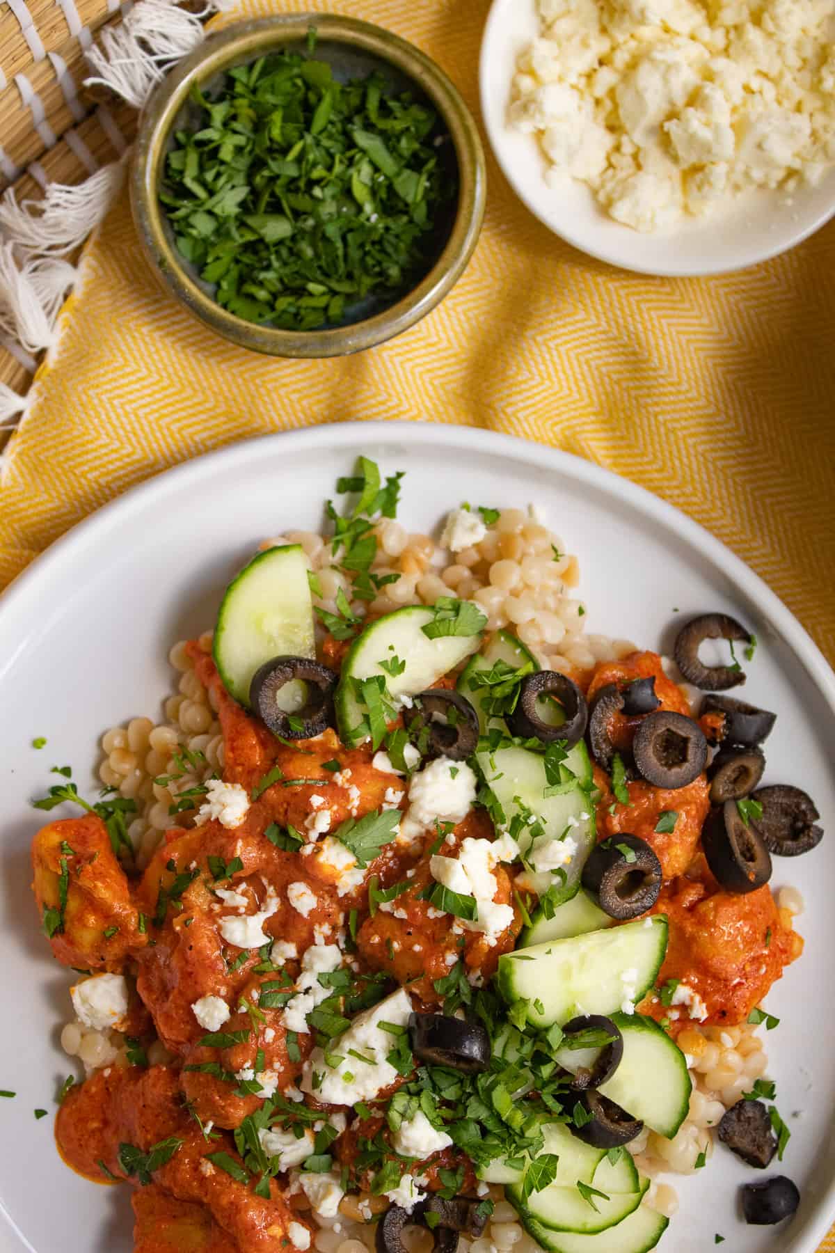 A white plate is piled with pearl cous cous, a vibrant orange sauce, and cucumber, feta cheese, parsley, and olive garnishes.