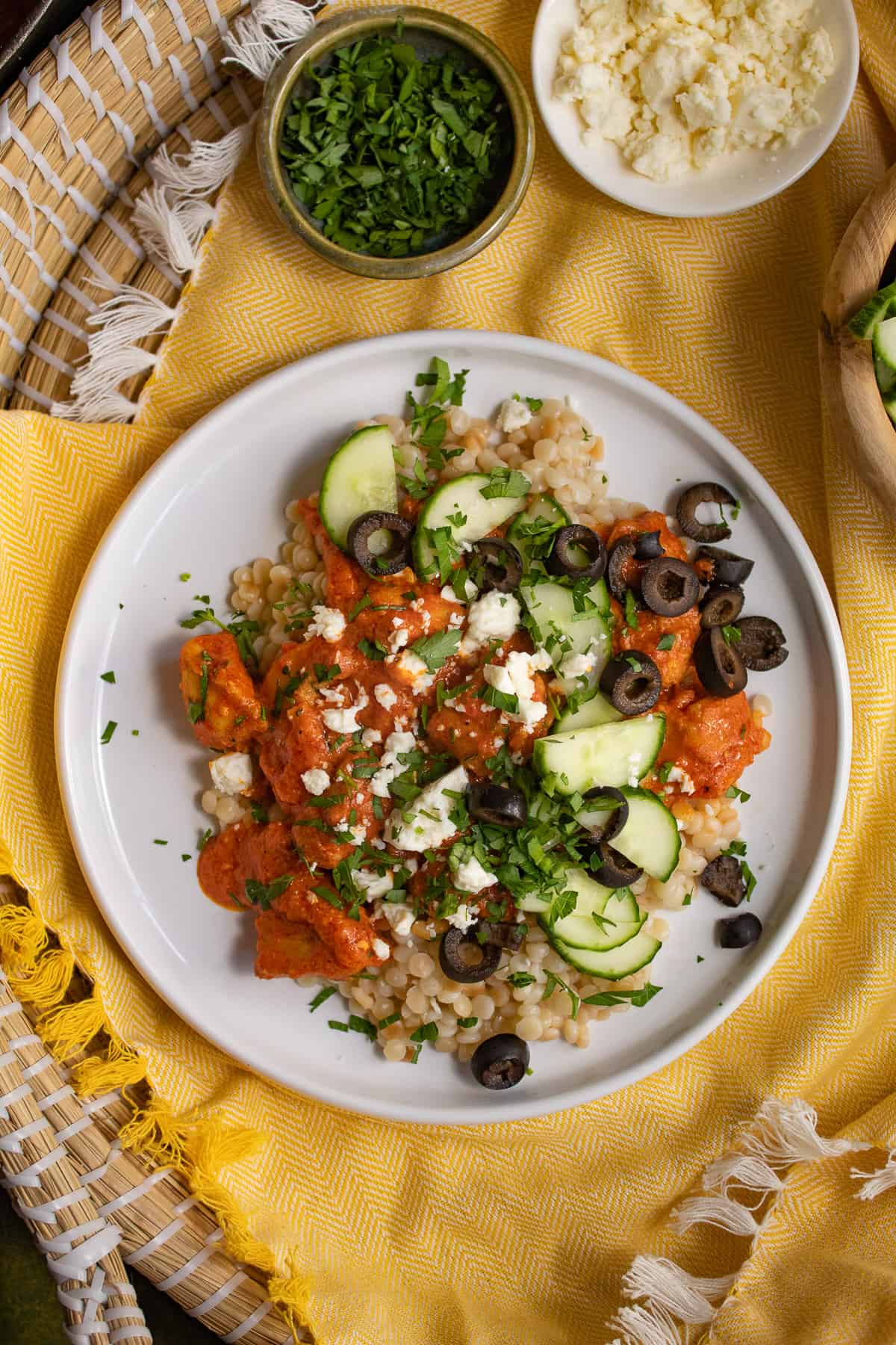 A white plate is piled with pearl cous cous, a vibrant orange sauce, and cucumber, feta cheese, parsley, and olive garnishes.