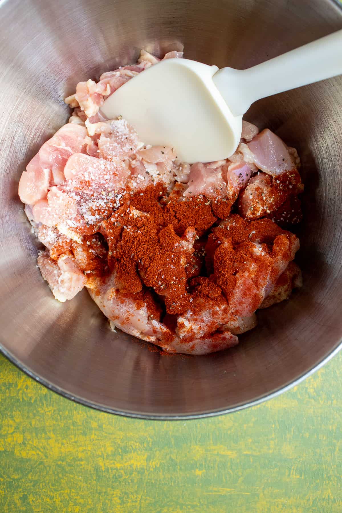 Chunks of chicken thighs in a silver bowl being seasoned with salt, pepper, and smoked paprika.