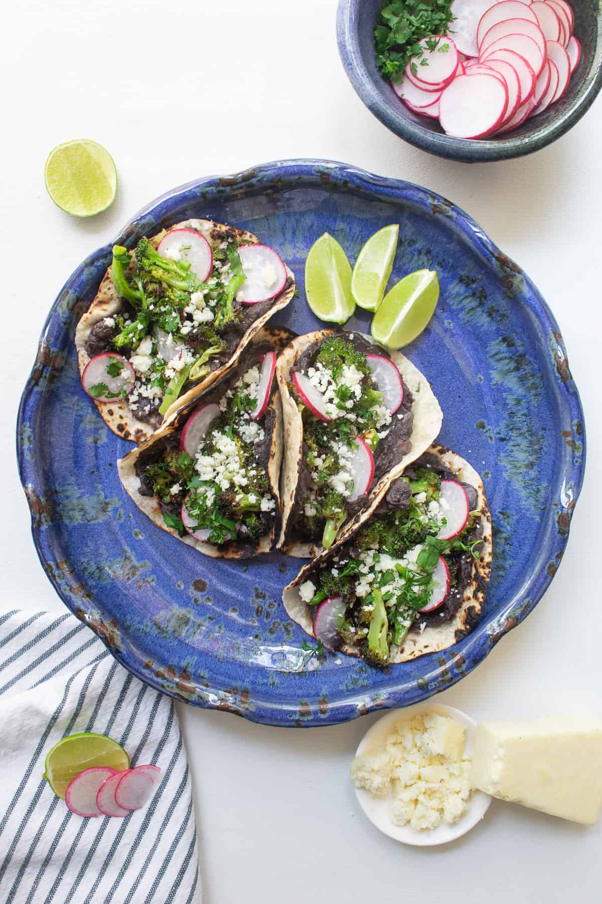 A blue plate is filled with bean and broccoli tacos in charred tortillas topped with radishes and crumbled cheese.