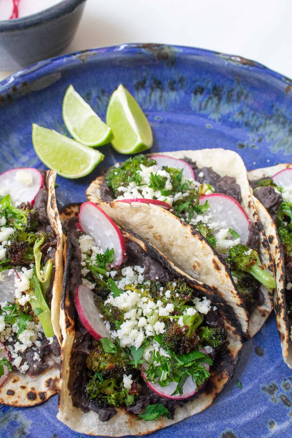 A blue plate is filled with bean and broccoli tacos in charred tortillas topped with radishes and crumbled cheese.