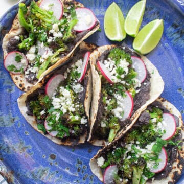 A blue plate is filled with bean and broccoli tacos in charred tortillas topped with radishes and crumbled cheese.