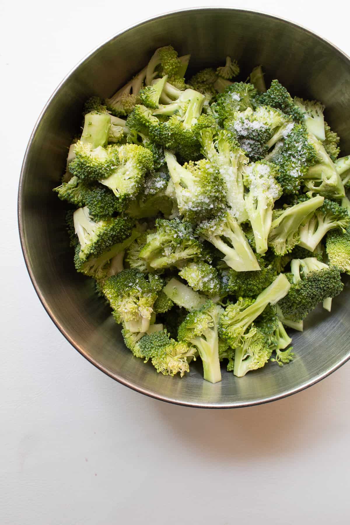 Broccoli florets in a metal bowl are sprinkled with salt.