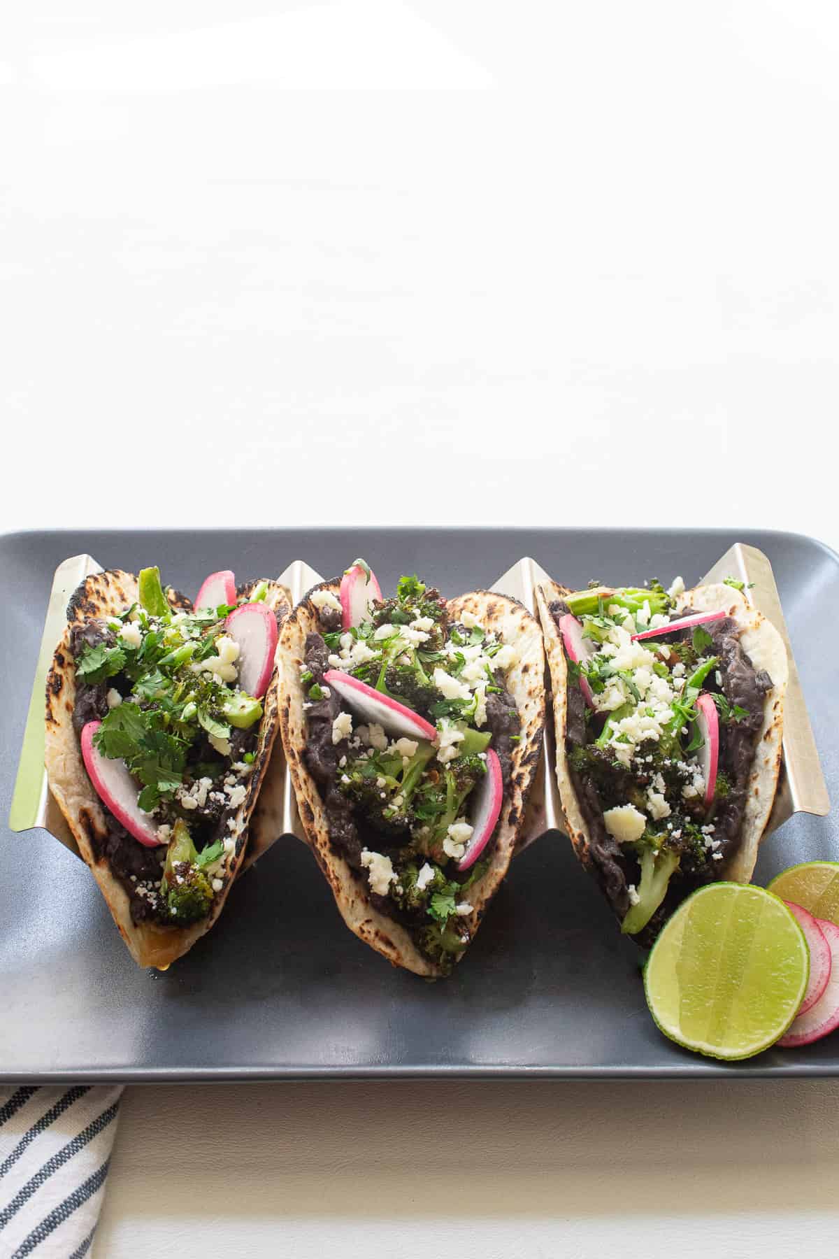 Three broccoli and black bean tacos sit in a taco holder on a gray plate.