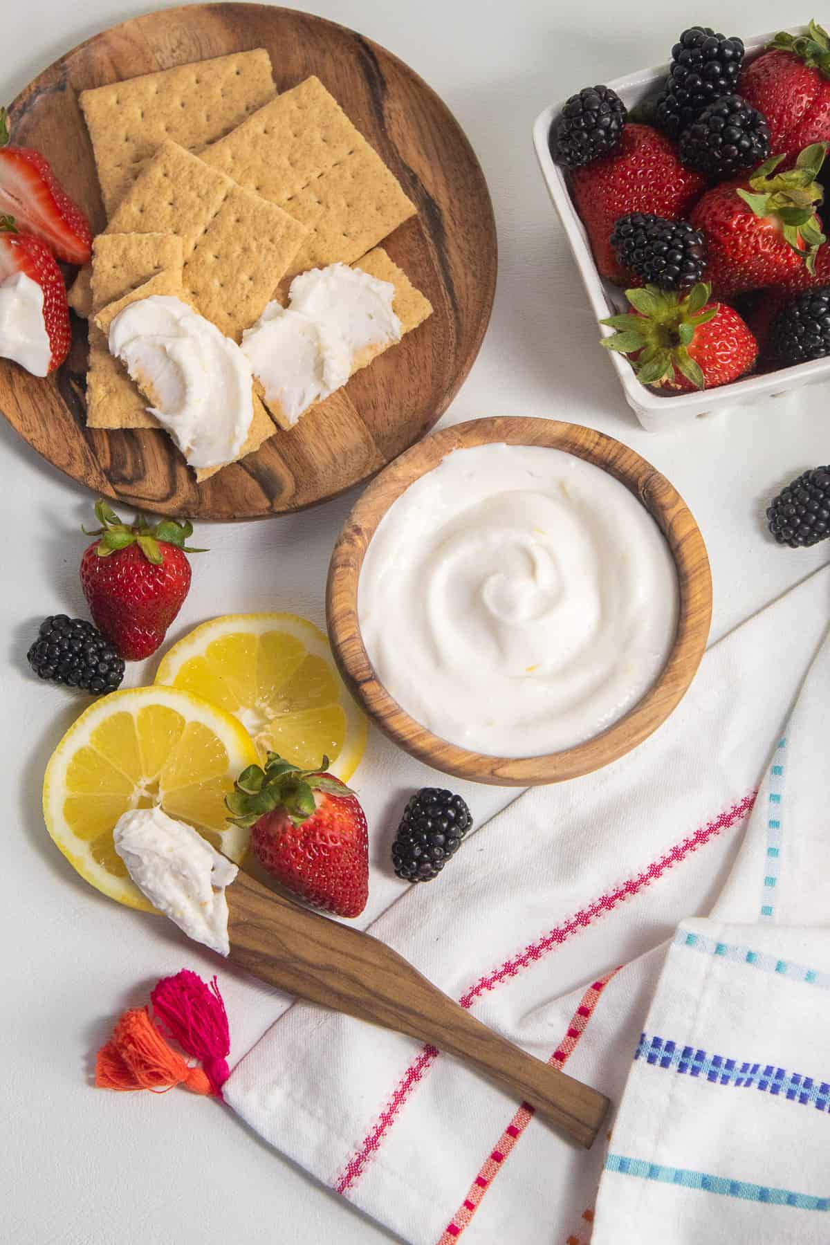 The fruit dip in a wooden bowl sits at the center of the image surrounded by fresh berries, lemon slices, and graham crackers.