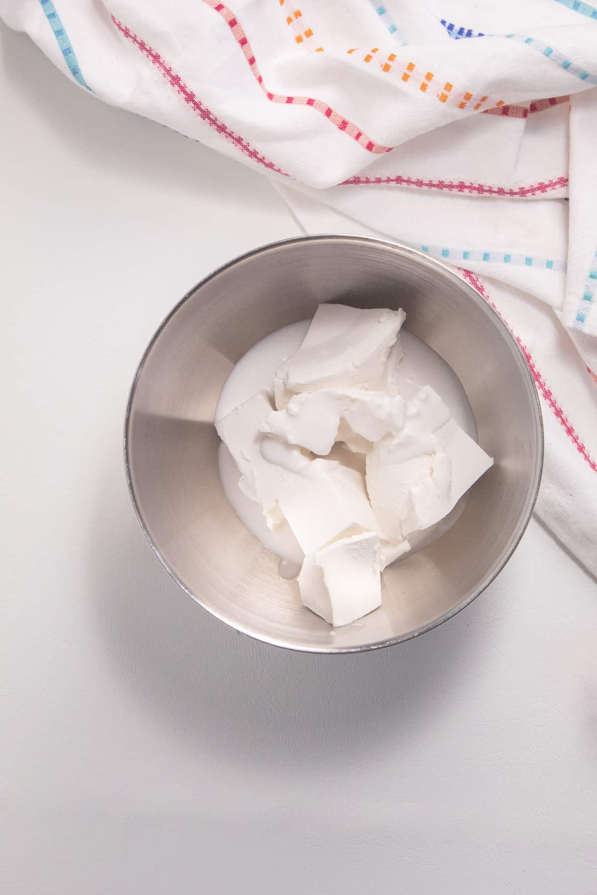 Cream cheese and heavy cream sit in the bottom of a silver bowl on a white surface.