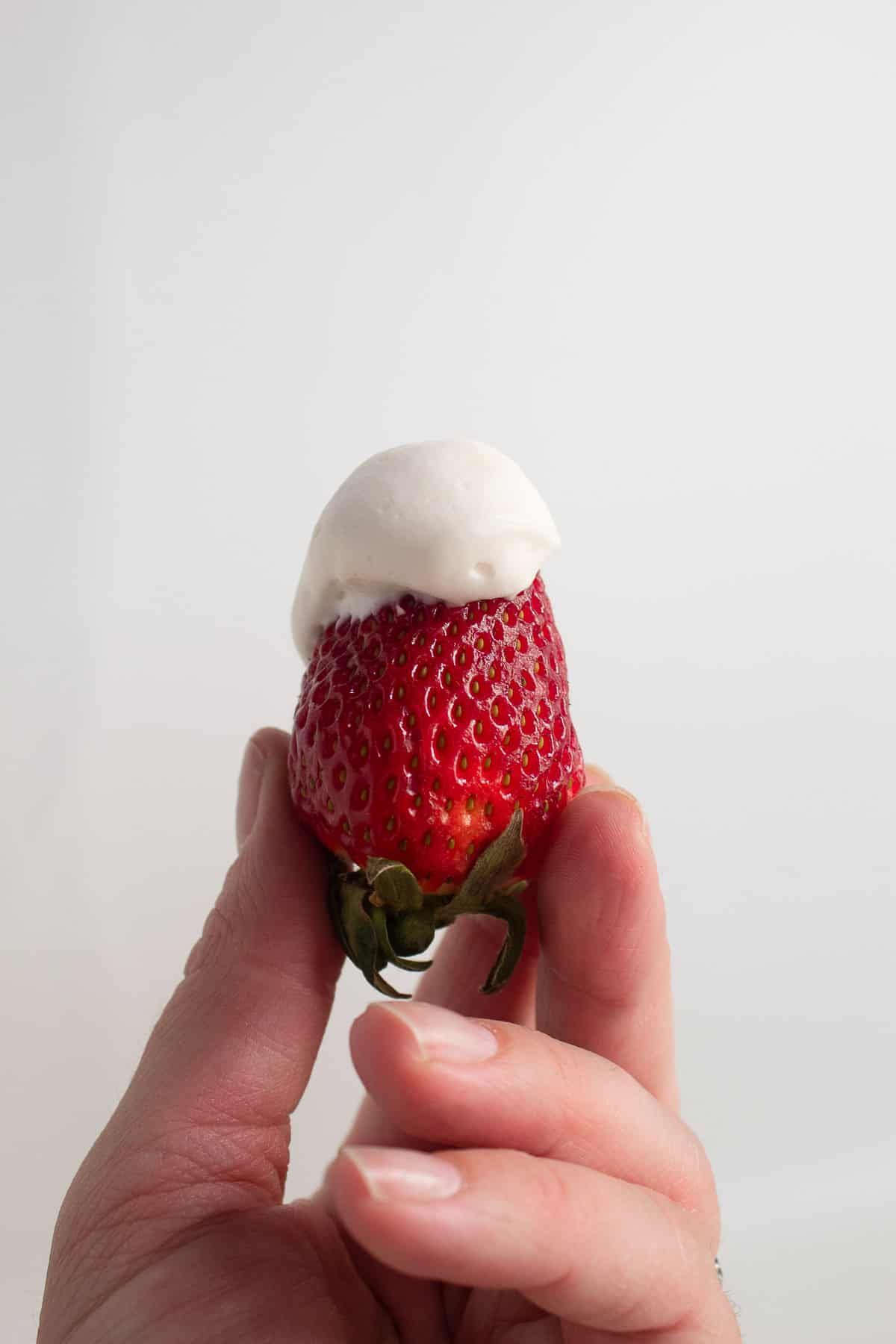 A red strawberry with white dip on the tip is held in the air by a hand.