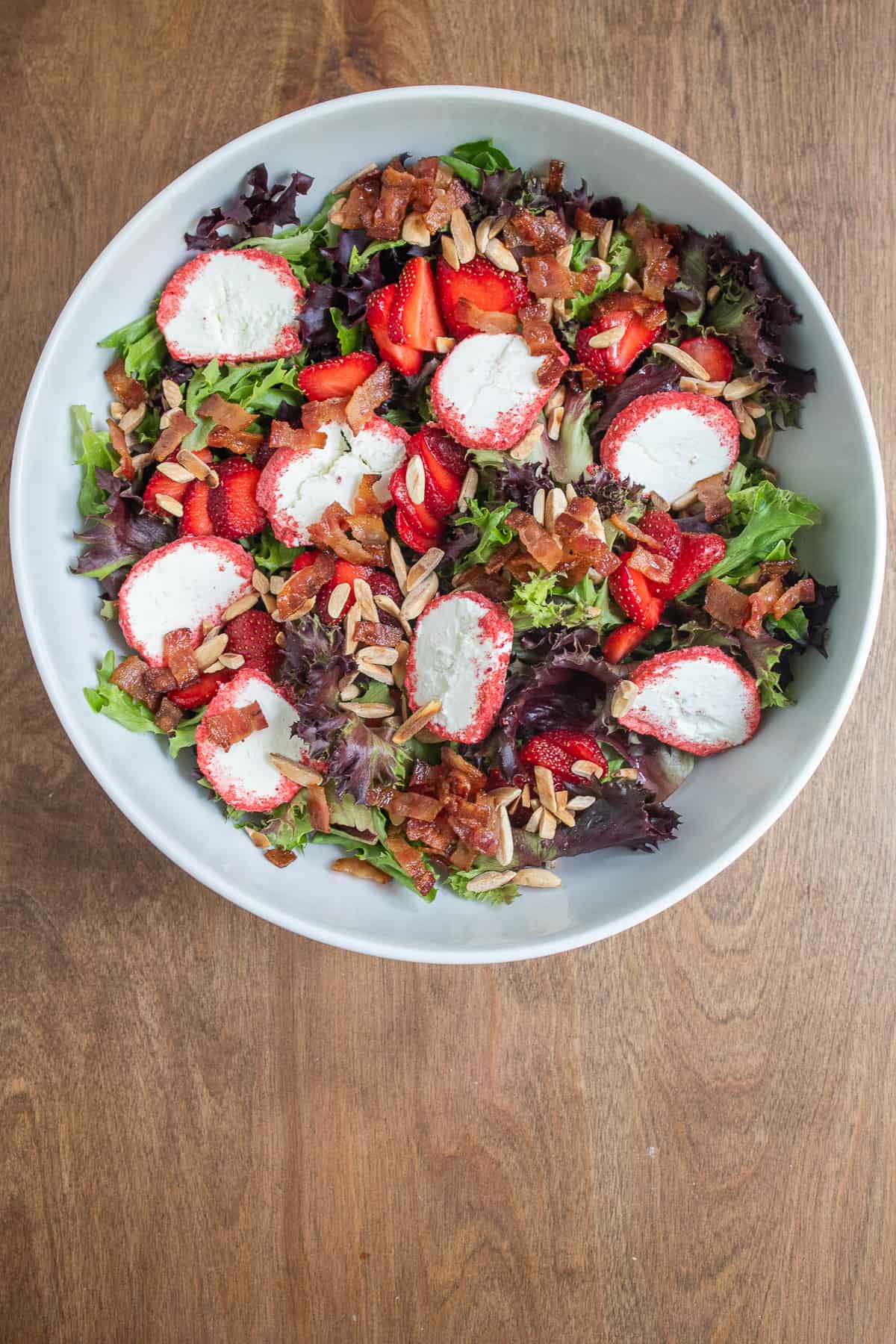 Mixed greens, almonds, bacon, strawberries, and strawberry-coated goat cheese are layered in a large white bowl.