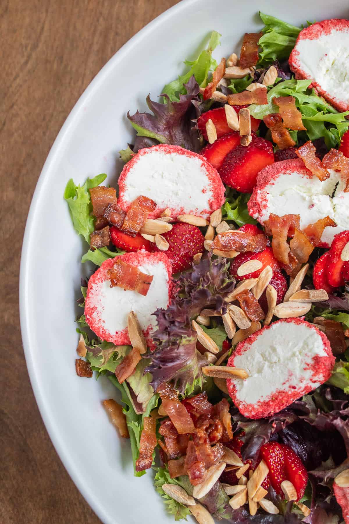 Mixed greens, almonds, bacon, strawberries, and strawberry-coated goat cheese are layered in a large white bowl.