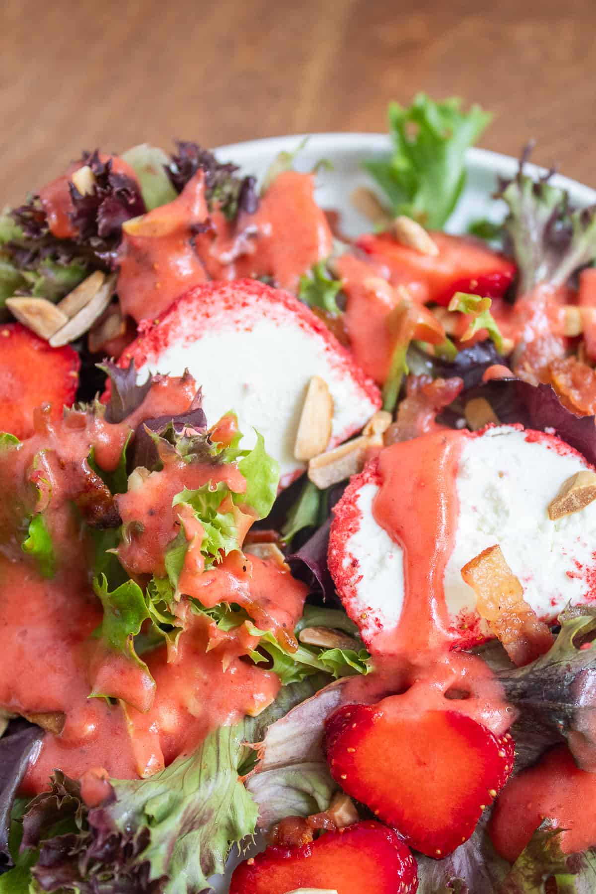 Detail image of mixed greens, fresh strawberries, almonds, bacon, goat cheese with pink coating, and a pink dressing.