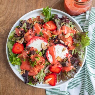 A white plate is piled with mixed greens, fresh strawberries, almonds, bacon, goat cheese with pink coating, and a pink dressing.