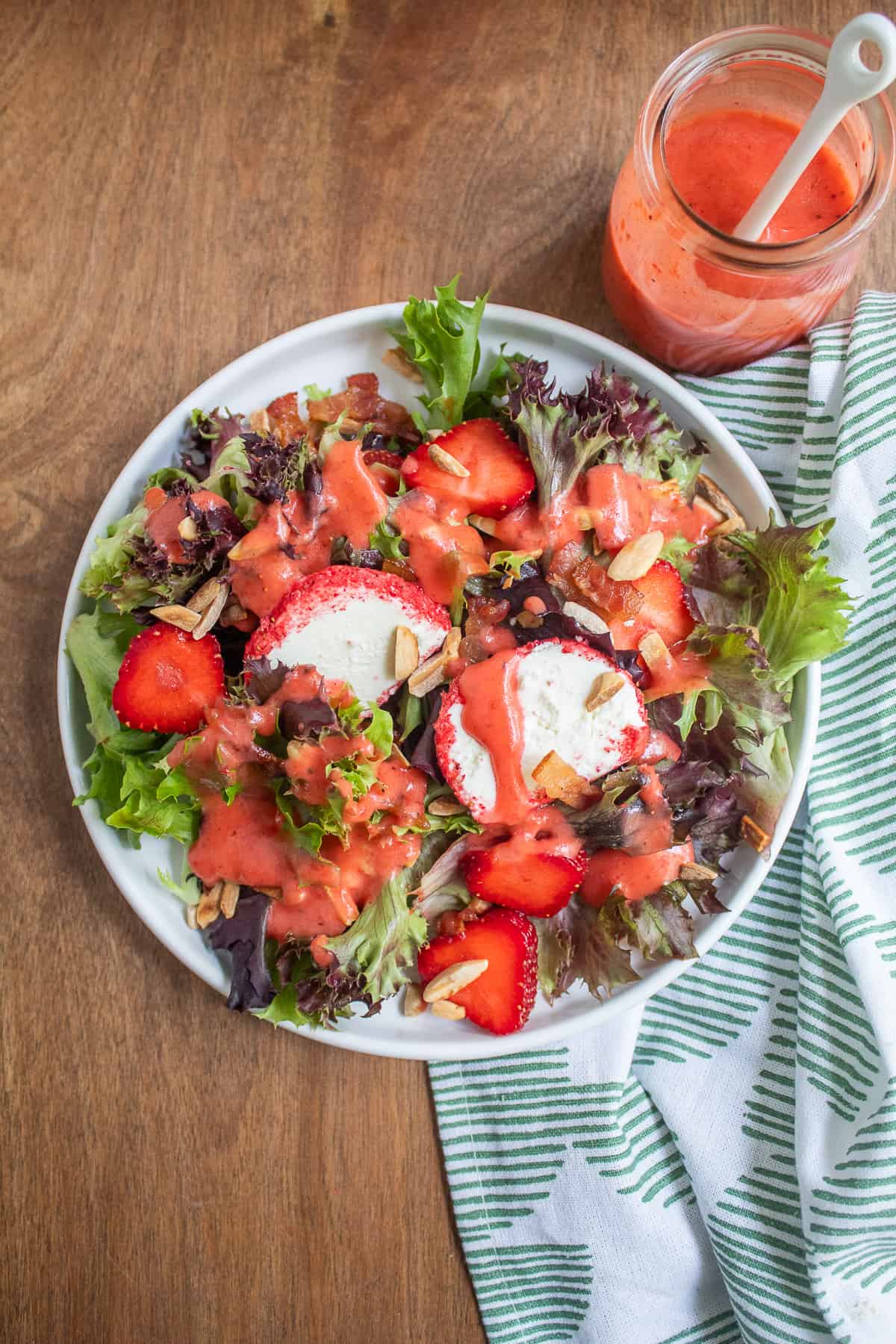 A white plate is piled with mixed greens, fresh strawberries, almonds, bacon, goat cheese with pink coating, and a pink dressing.