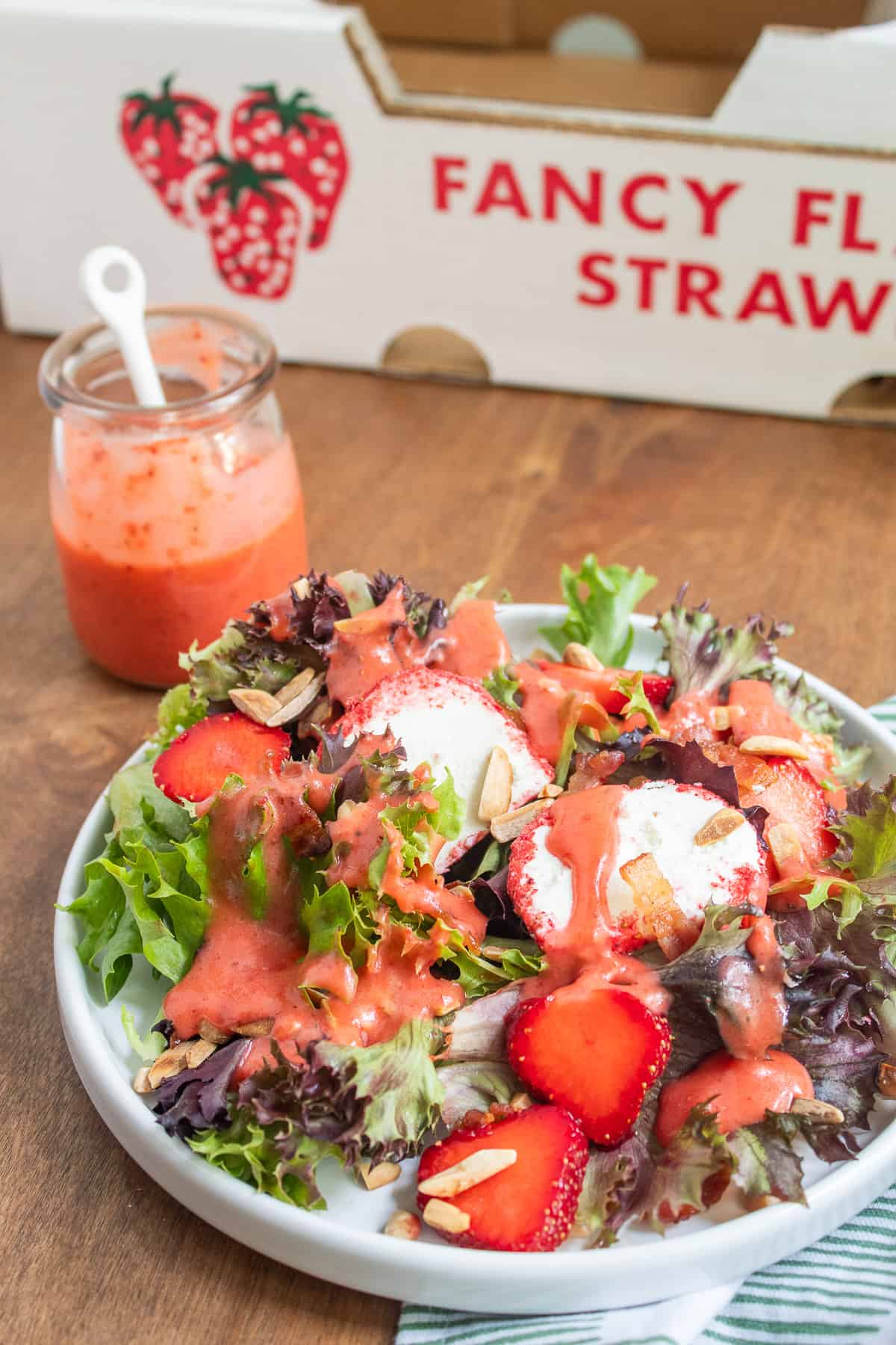 A white plate is piled with mixed greens, fresh strawberries, almonds, bacon, goat cheese with pink coating, and a pink dressing.