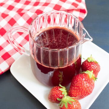 A small pitcher of red syrup sits on a white plate next to fresh strawberries.