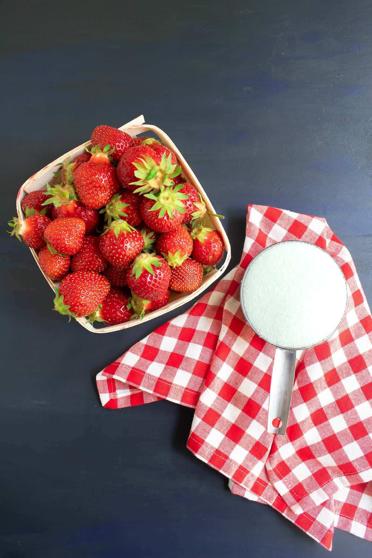 Fresh strawberries sit in a wooden basket alongside a cup of granulated sugar on a red and white checked napkin on a blue surface.
