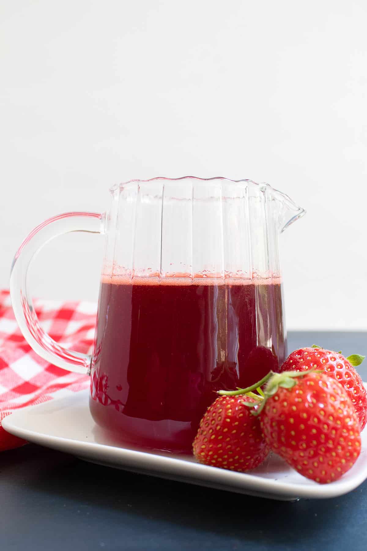 A small pitcher of red syrup sits on a white plate next to fresh strawberries.