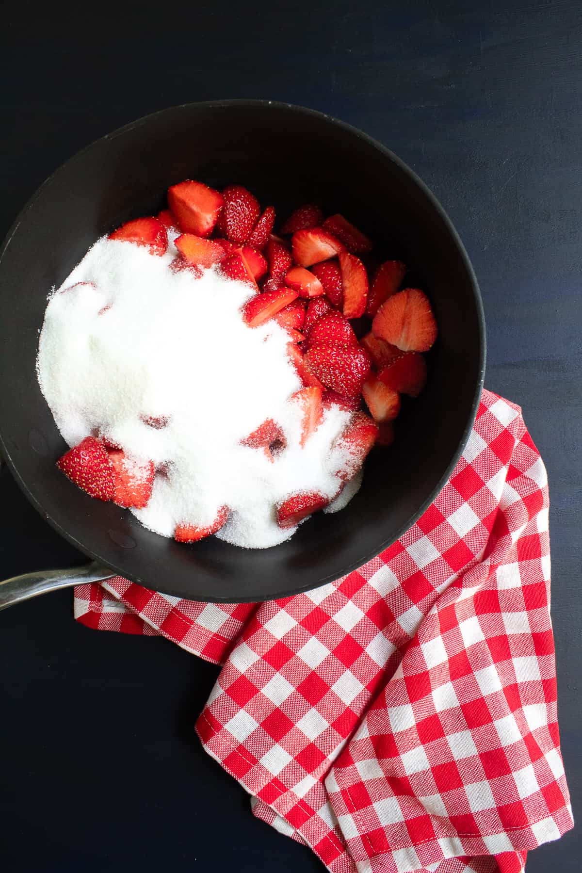Strawberries and granulated sugar are piled into a black saucepan.