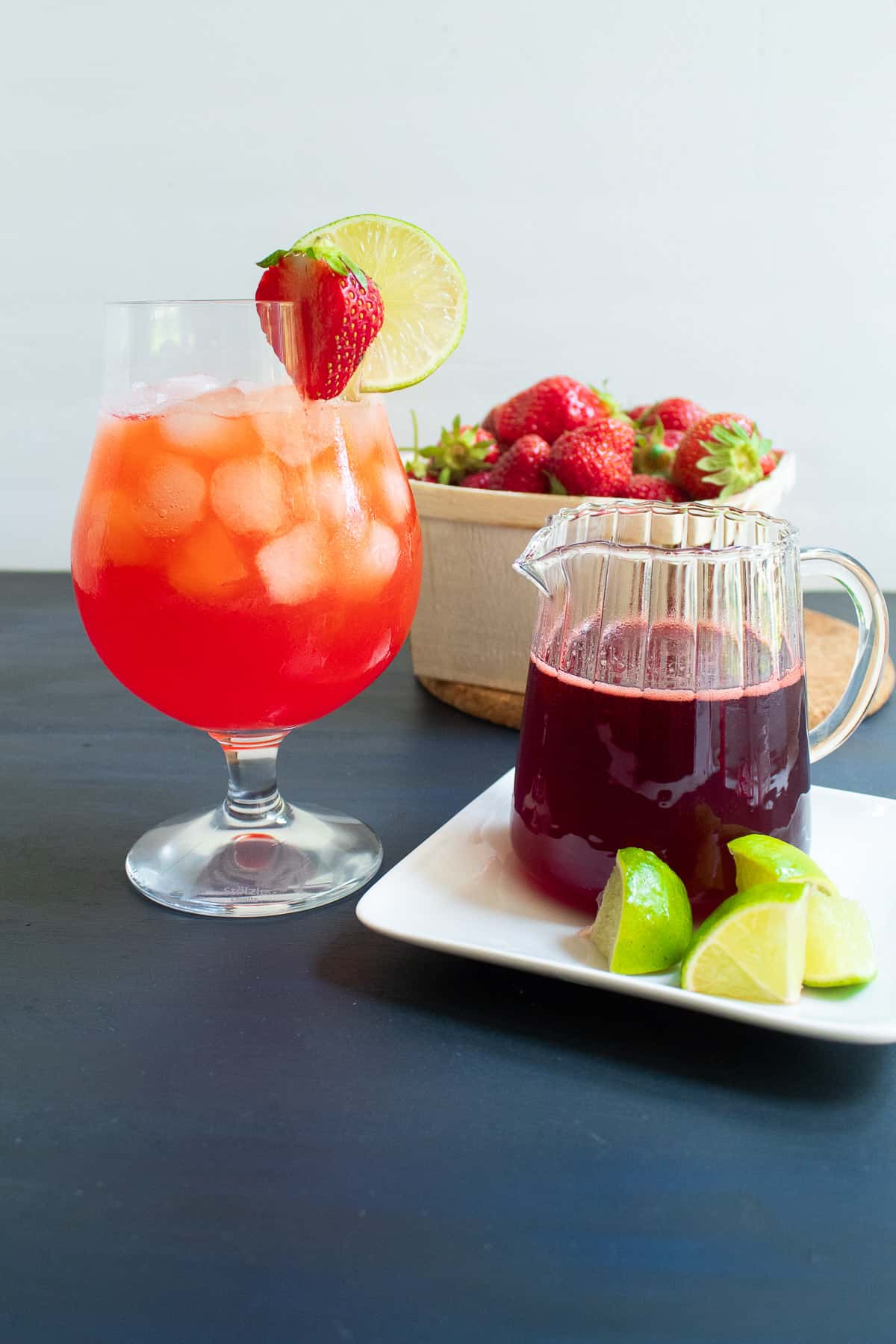 A large glass of strawberry limeade is garnished with a fresh strawberry and a lime wheel sits next to a pitcher of strawberry syrup and lime wedges.