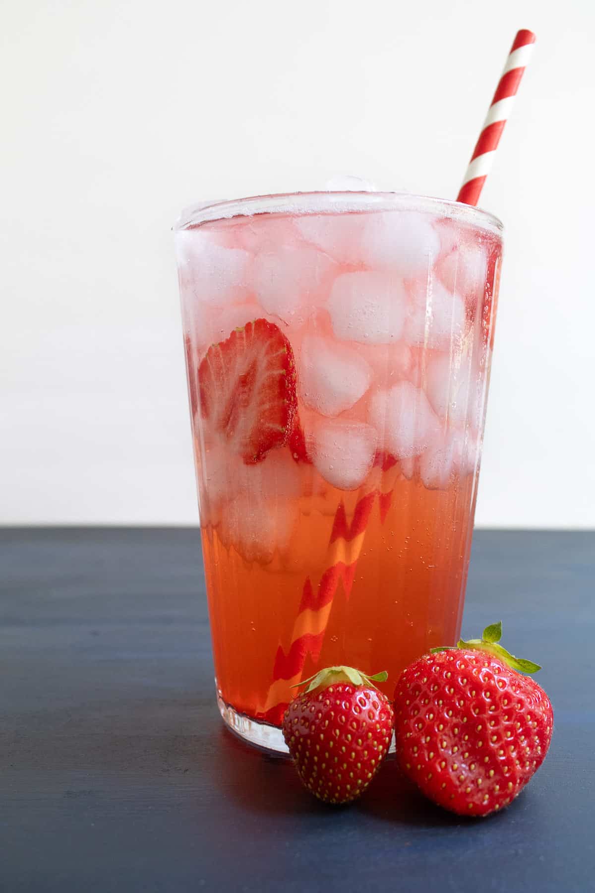 A tall glass of a red beverage with ice and a red and white striped straw sits on a blue surface alongside two fresh strawberries.