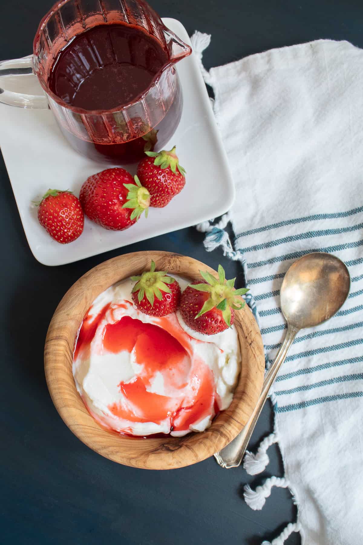 A wood bowl with swirled yogurt, strawberries, and red syrup sits on a blue surface near a pitcher of dark red syrup.