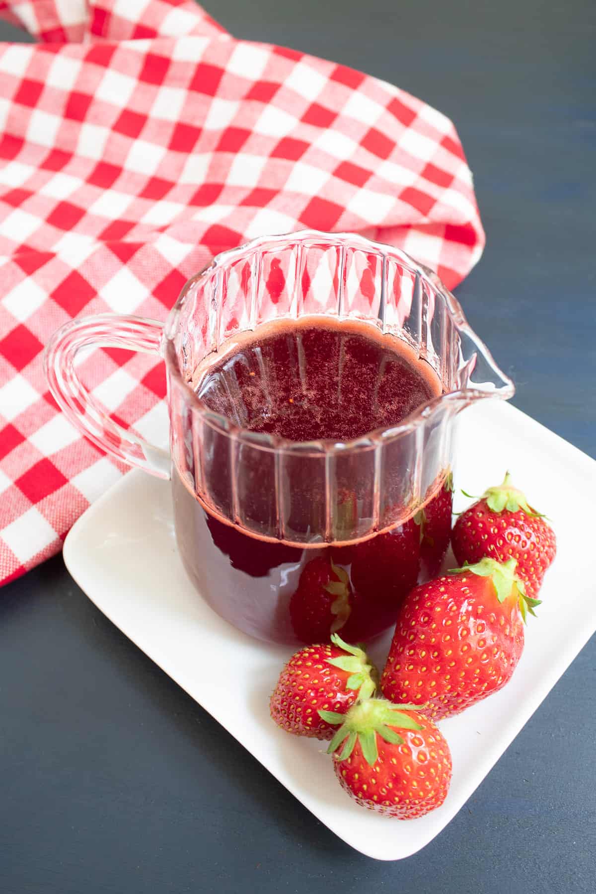 A pitcher of red syrup sits on a white plate near a red and white checked napkin.