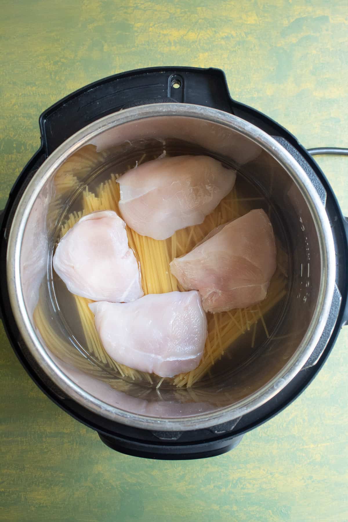 The uncooked spaghetti is covered with water and the halved chicken breasts are sitting atop the spaghetti in the Instant Pot insert.