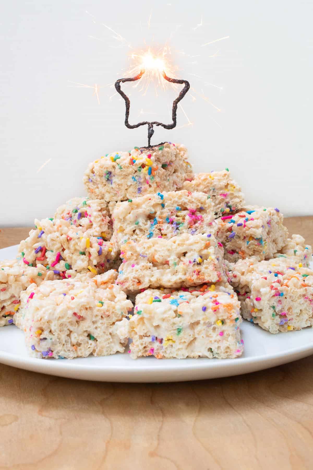 A star-shaped sparkler is throwing sparks atop a pile of treats on a white plate.