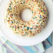 A rice krispie cake molded into a ring sits on a white cake platter over a multicolored stripe towel.
