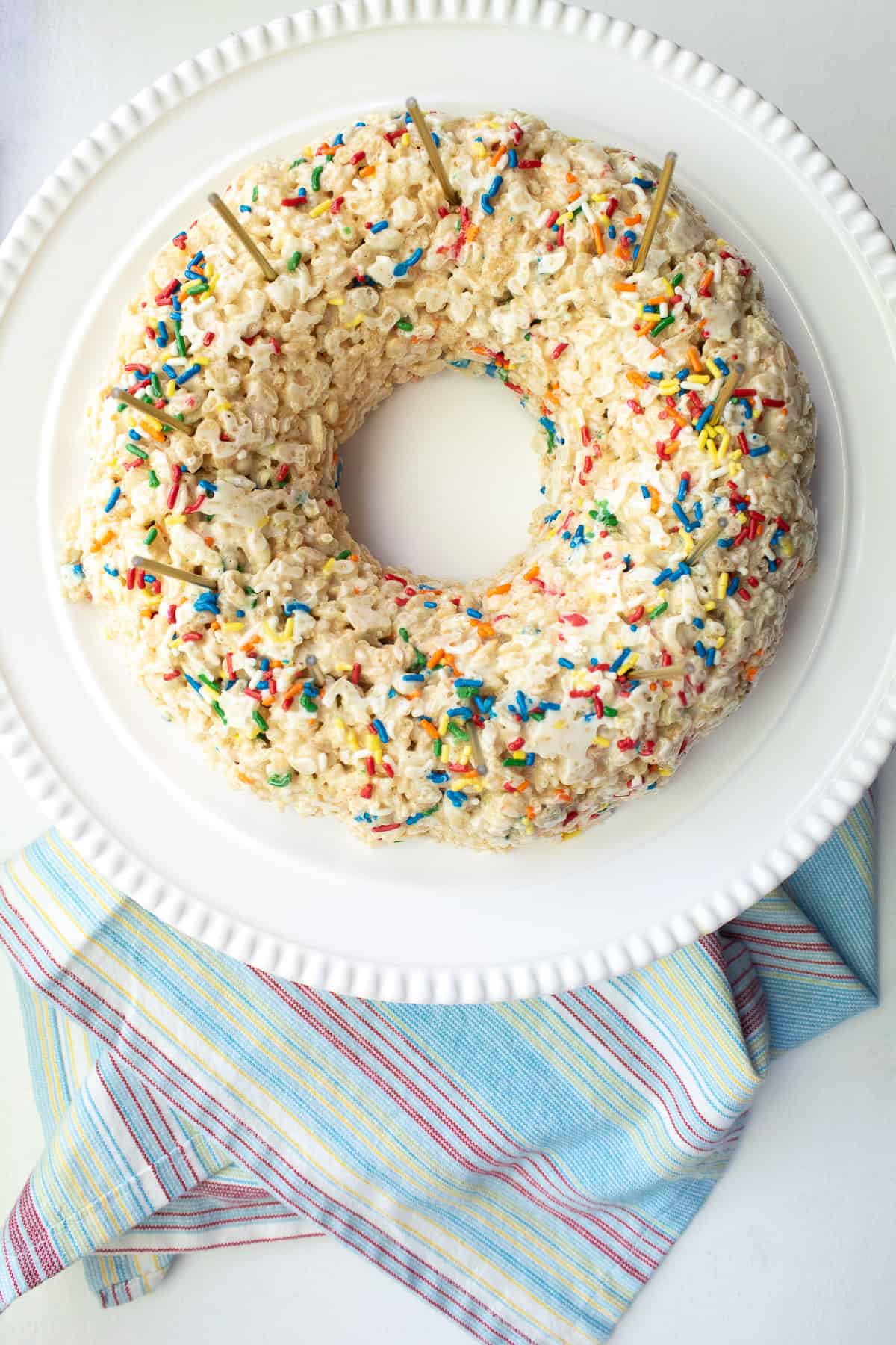 A rice krispie cake molded into a ring sits on a white cake platter over a multicolored stripe towel.