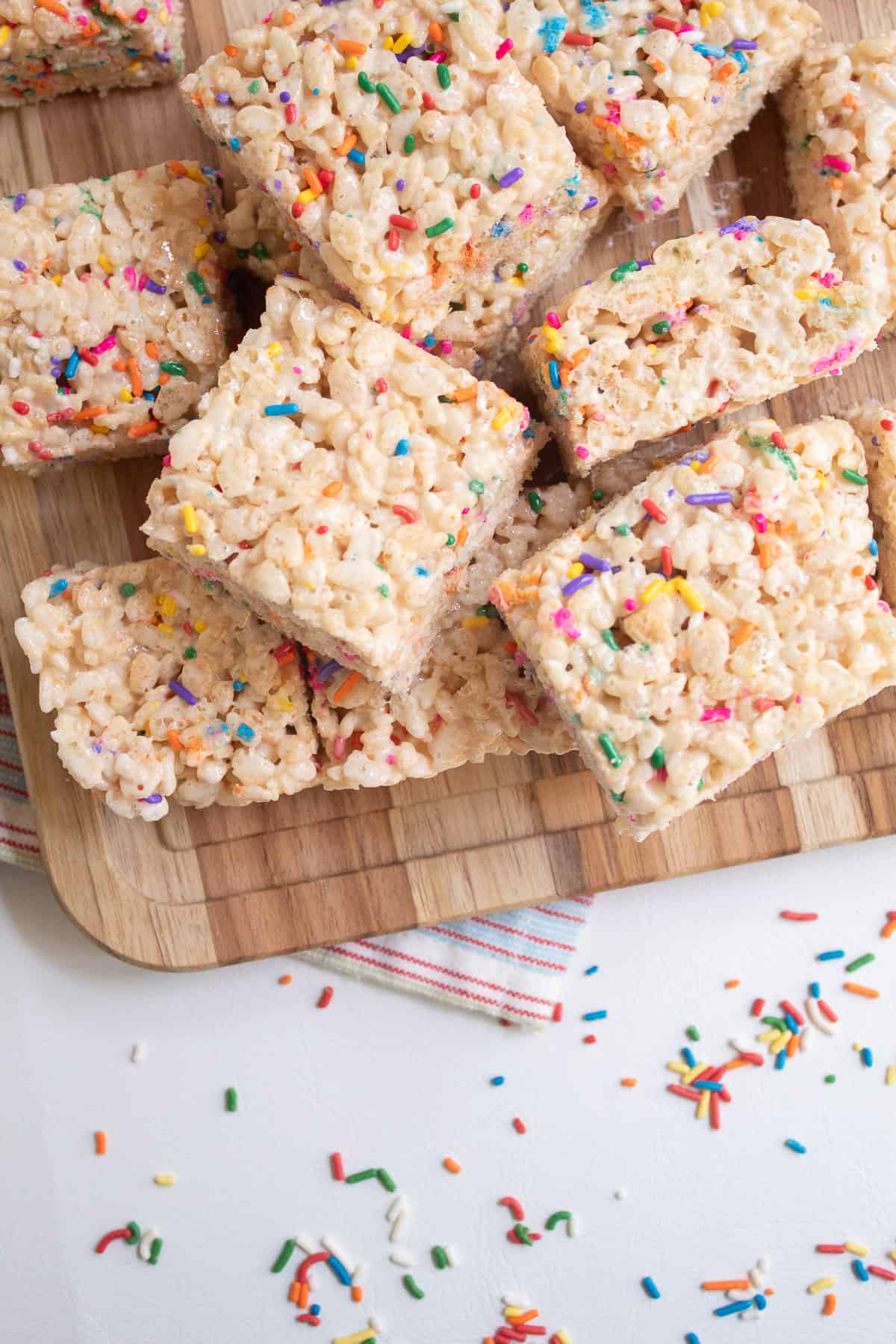 Finished treat squares are stacked on a wooden cutting board over a surface scattered with sprinkles.