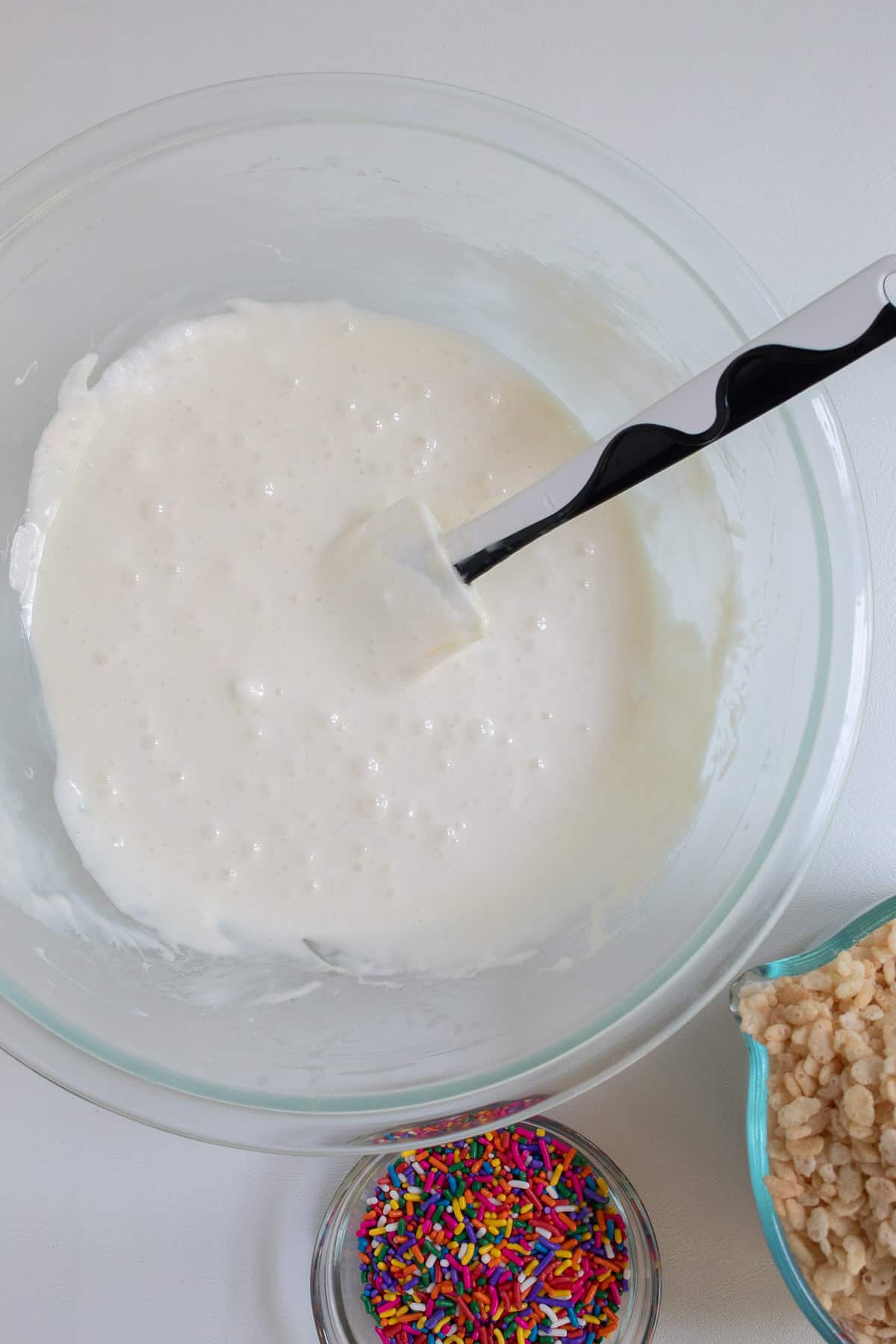 The melted marshmallow mixture in a clear glass bowl next to smaller bowls of sprinkles and crisp rice cereal.