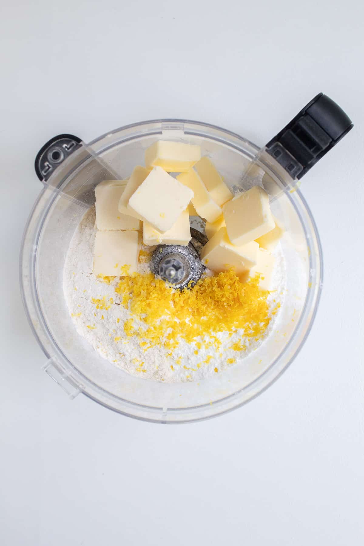 Powdered sugar, flour, lemon zest, and butter sit in the bowl of a food processor.