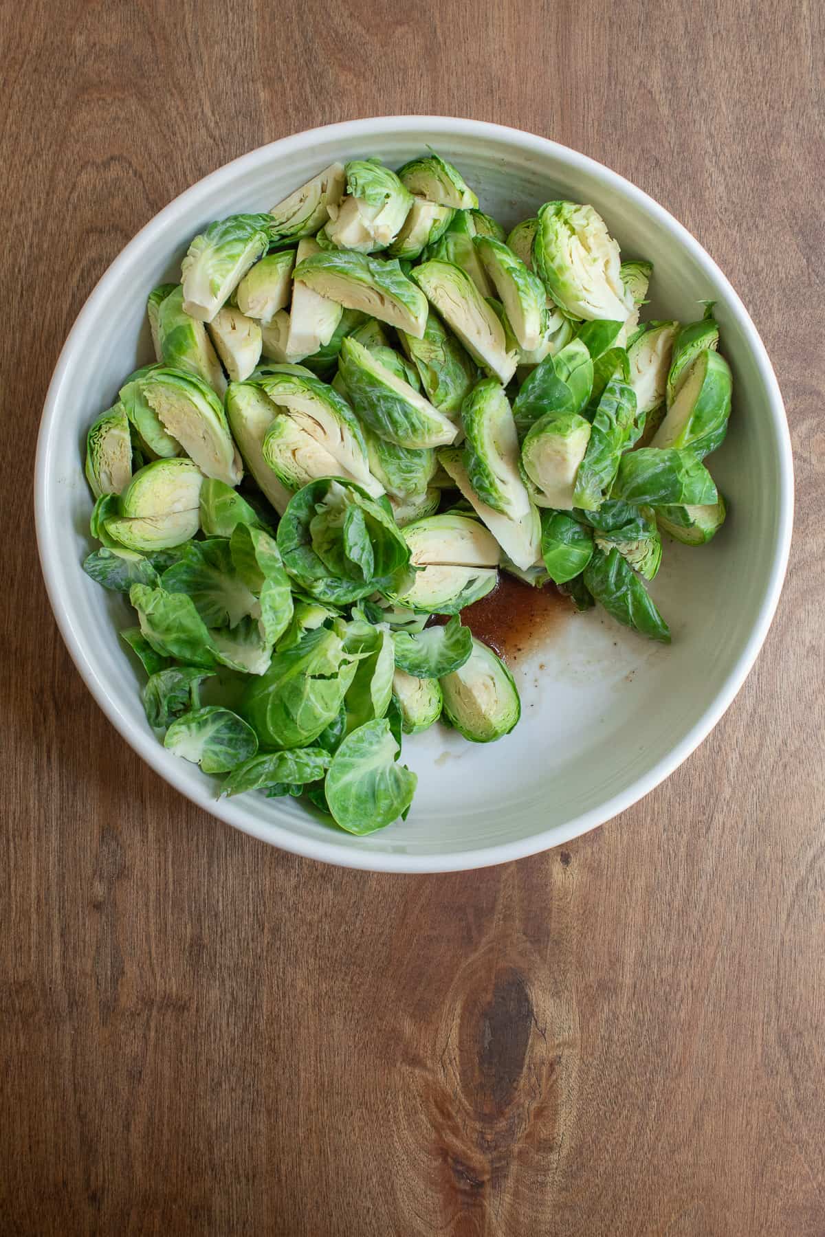 The trimmed brussel sprouts sit in a large white bowl with the glaze mixture at the bottom.