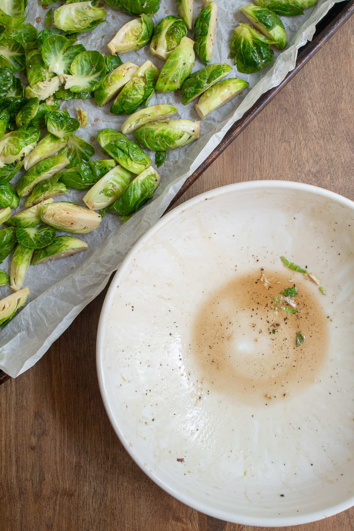 The bowl used for tossing the sprouts with the glaze has some remaining glaze in it. It sits next to the sheet pan loaded with the brussel sprouts.
