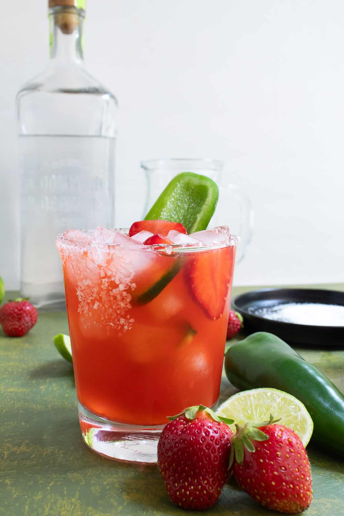 A rocks glass with a salted rim holds a red liquid and is garnished with a sliced strawberry and a slice of jalapeno. In the background sits a tall clear glass bottle containing a clear liquid.