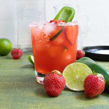 A rocks glass with a salted rim holds a red liquid and is garnished with a sliced strawberry and a slice of jalapeno. In fornt of the glass are two fresh strawberries, a sliced lime, and a fresh jalapeno.